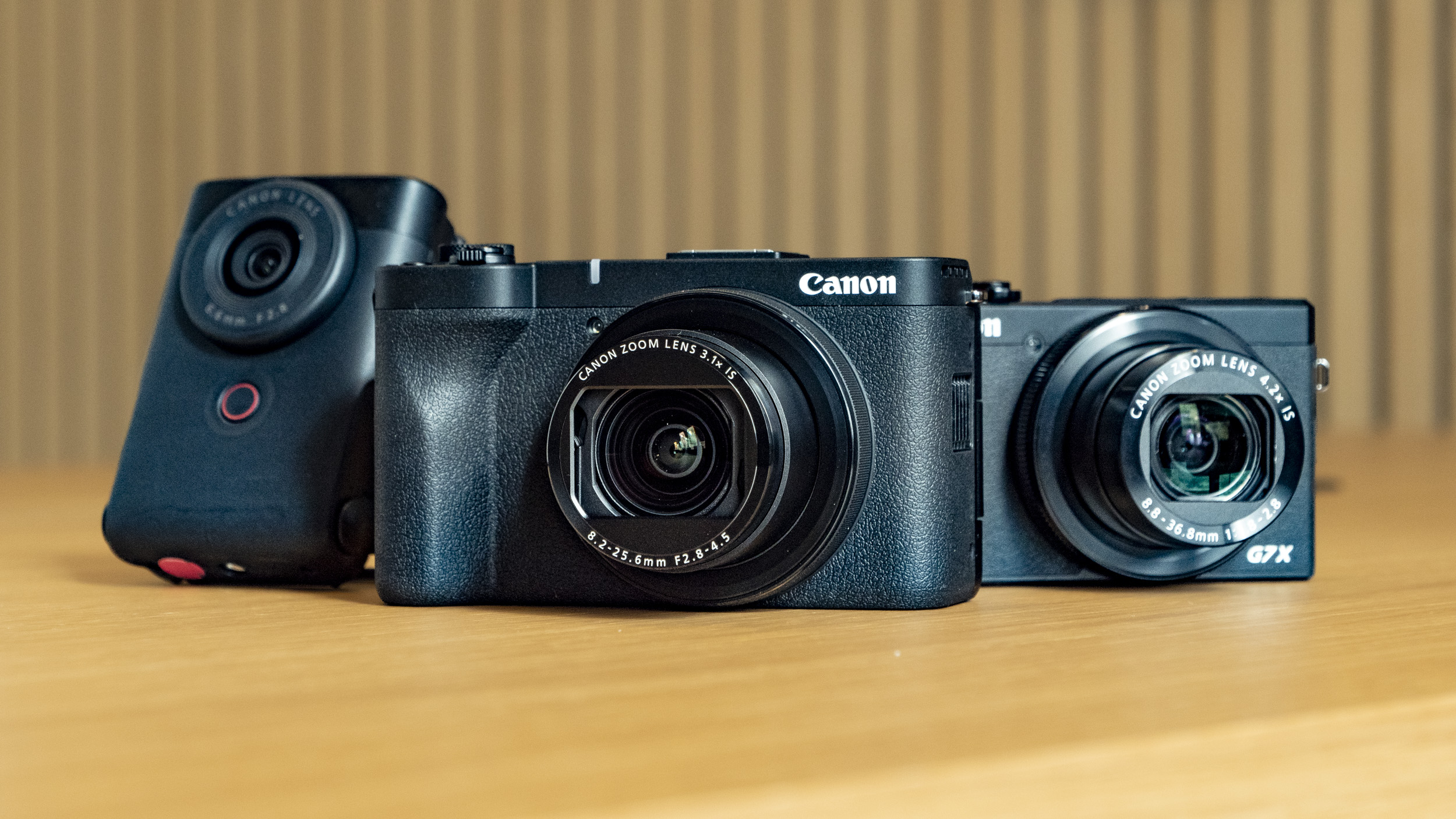 Canon PowerShot V1 compact vlogging camera on a wooden desk alongside the PowerShot G7X Mark III and PowerShot V10
