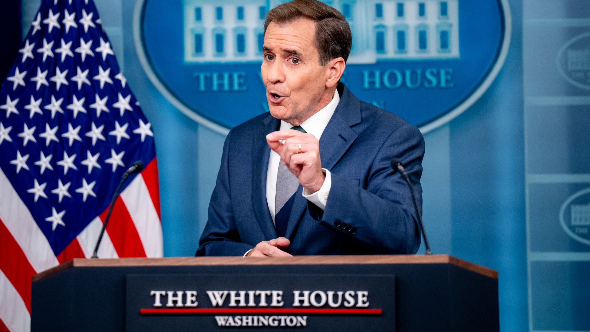 a man with short hair in a blue suit speaks into microphones at a lectern featuring a sign reading "the white house" in front of an american flag