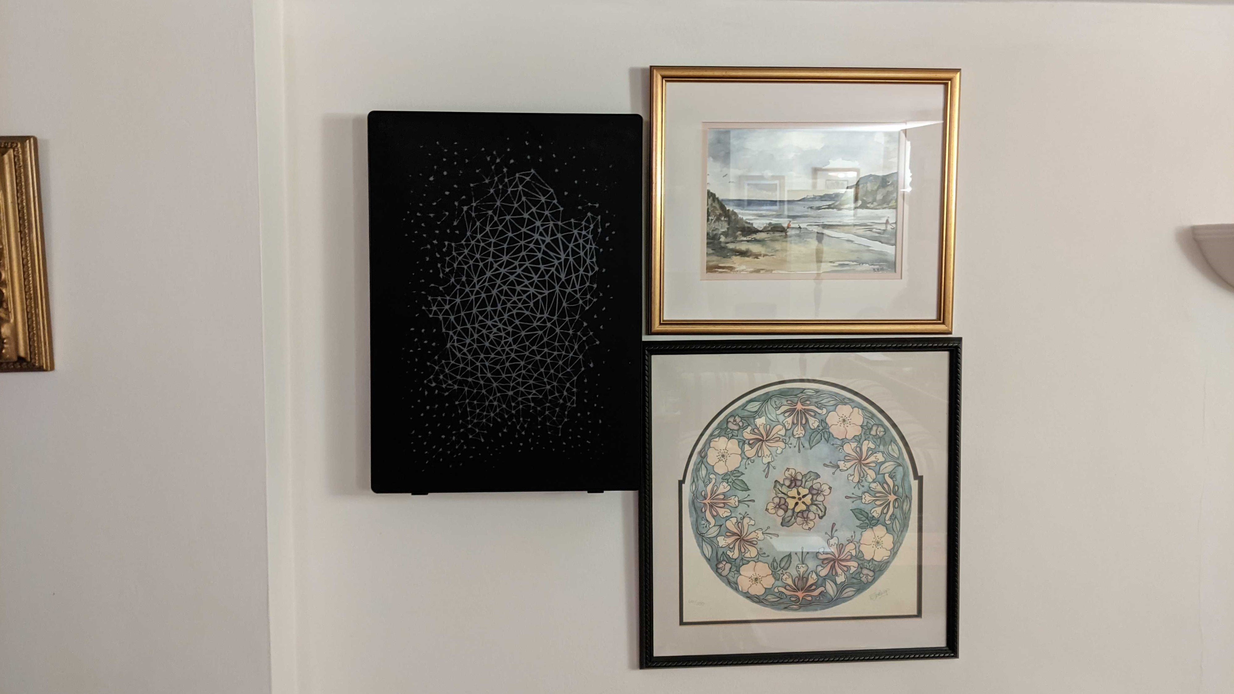The Symfonisk Picture Frame hanging on a wall next to two framed pictures a beach and a floral pattern