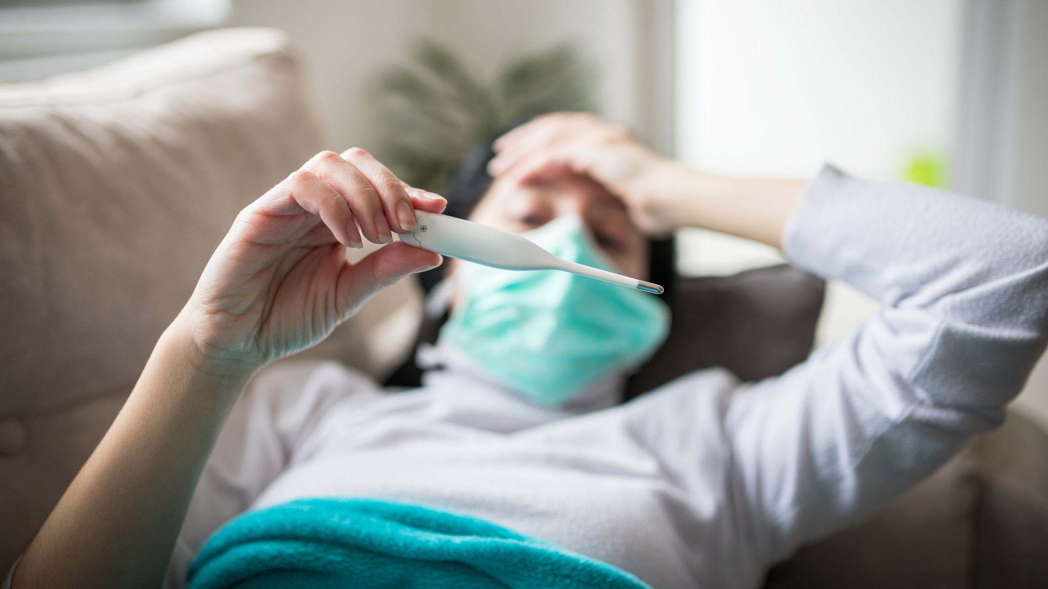 Sick person wearing a surgical mask and laying on a couch. They're holding their head with one hand and holding up a thermometer with the other, checking for fever