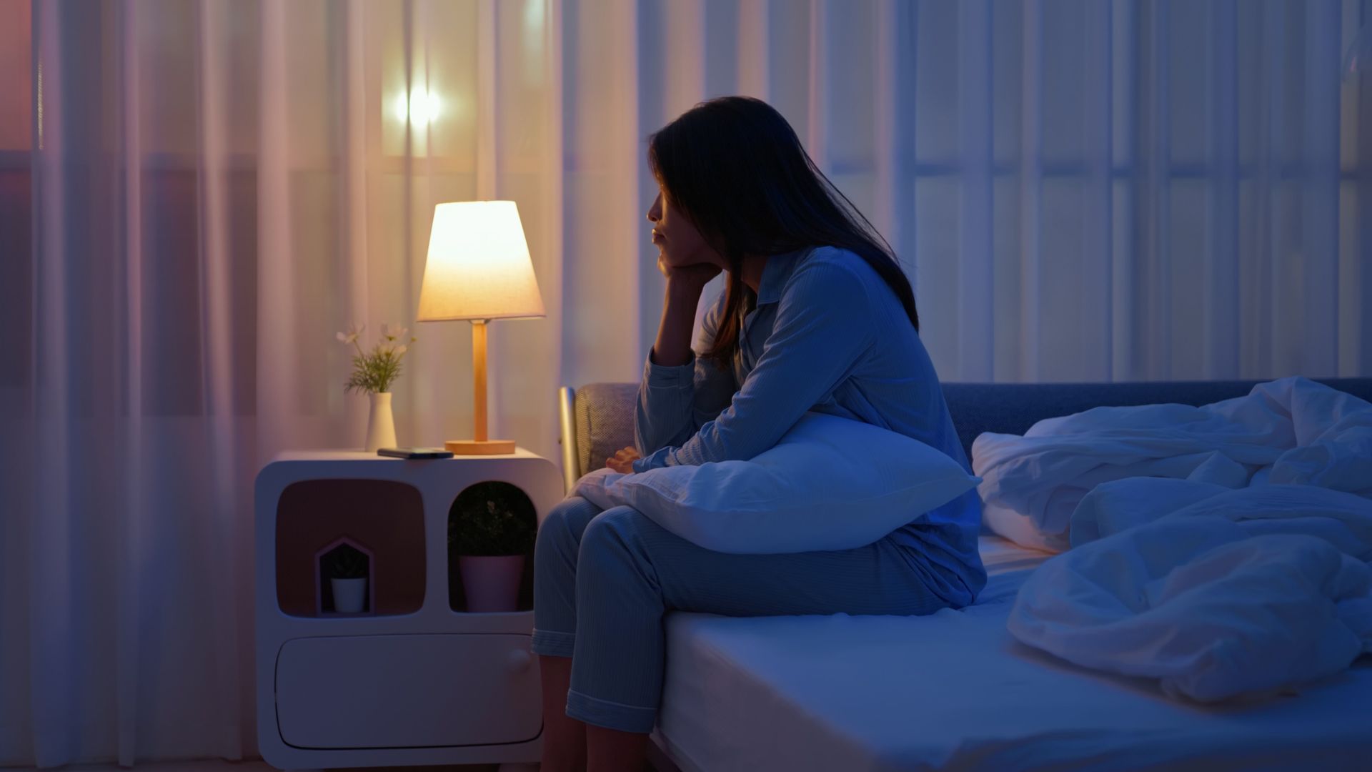 A young woman sits at the edge of her bed in a dimly lit bedroom as she can&#039;t sleep due to worry. 